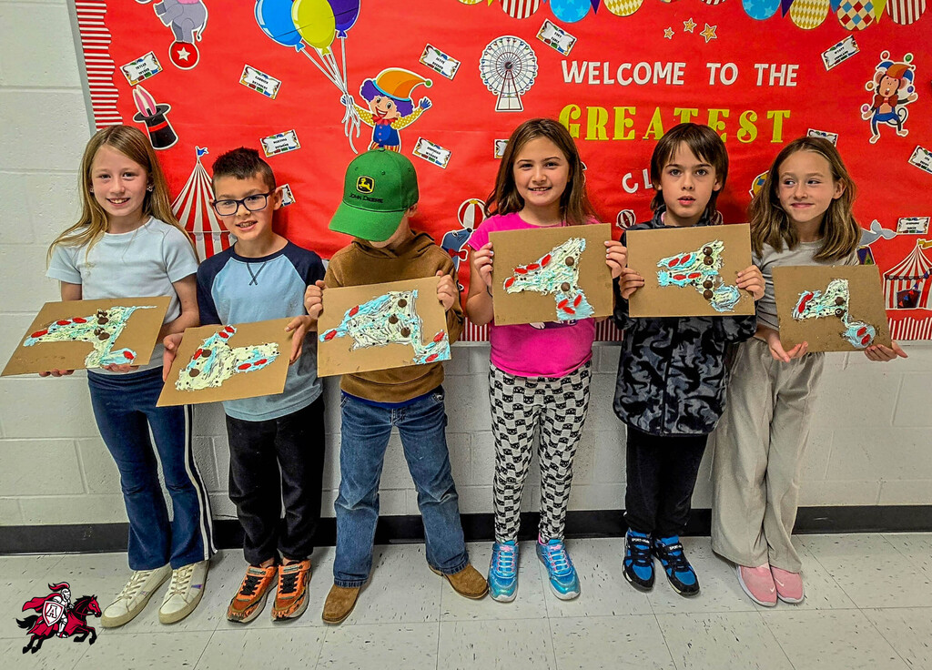 Six children in a classroom hold artwork. The backdrop is a red banner with the words "Welcome to the Greatest."