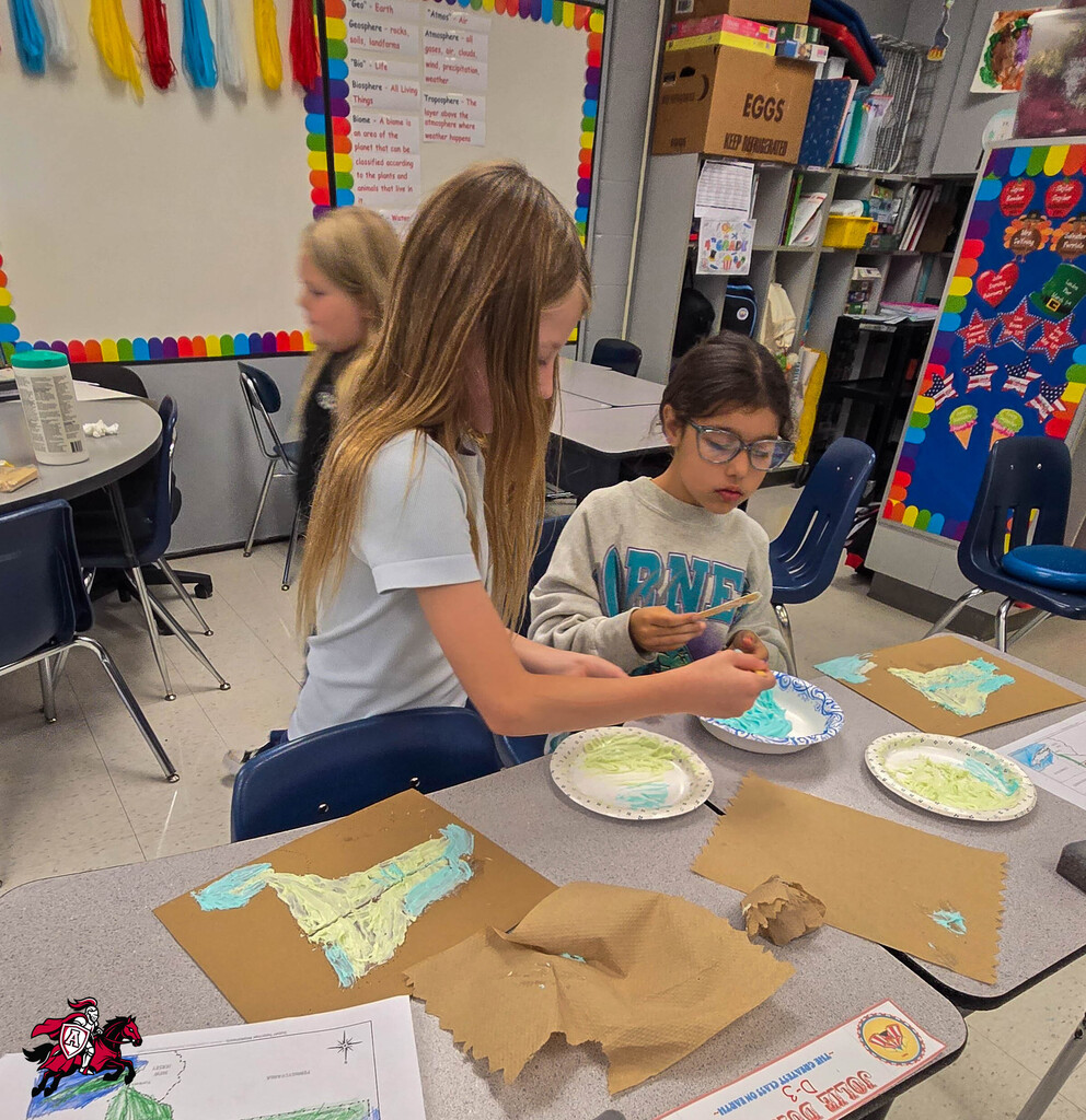 Two children at a table in a classroom working on crafts. One child is holding a paintbrush.