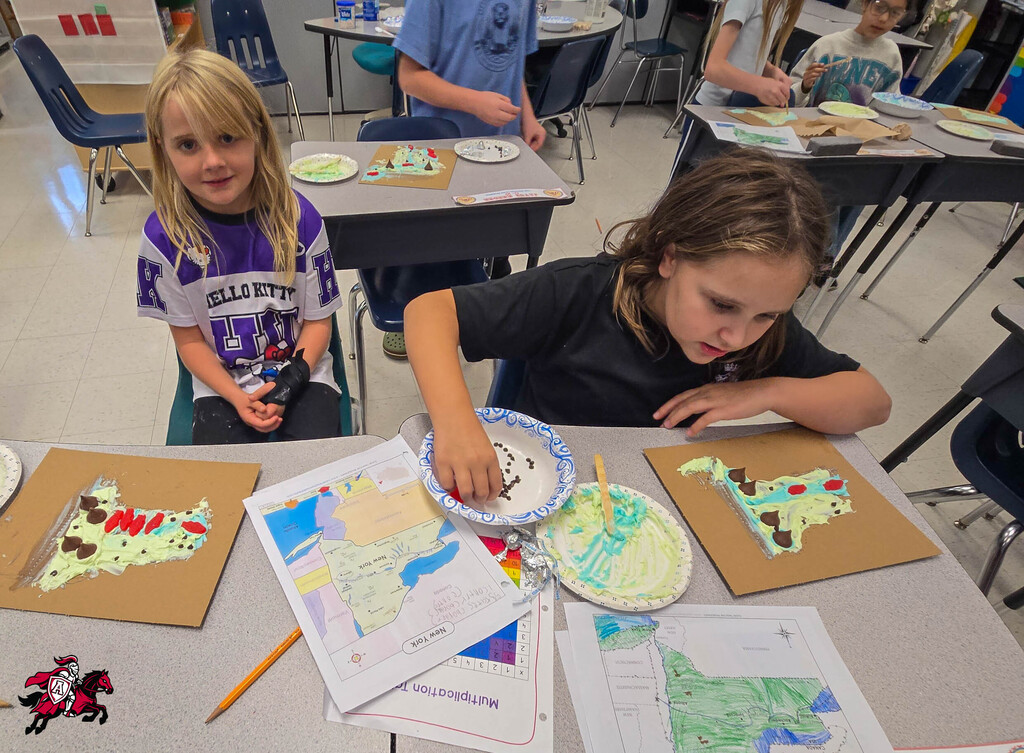 Two girls in a classroom, one with blonde hair and a purple shirt, the other with brown hair and a black shirt, are creating art.