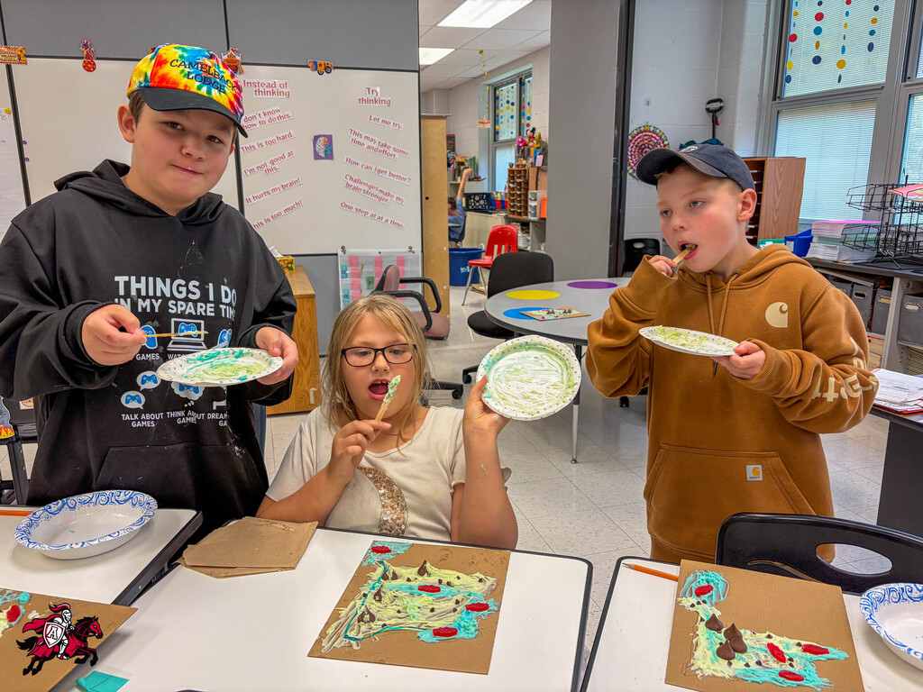 Three kids hold plates in a classroom, one with a chocolate-covered toothbrush. Behind them, tables and chairs are visible.