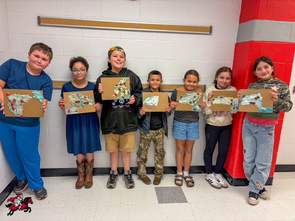 Seven children in a hallway, holding up artwork. A red and gray pillar behind them.