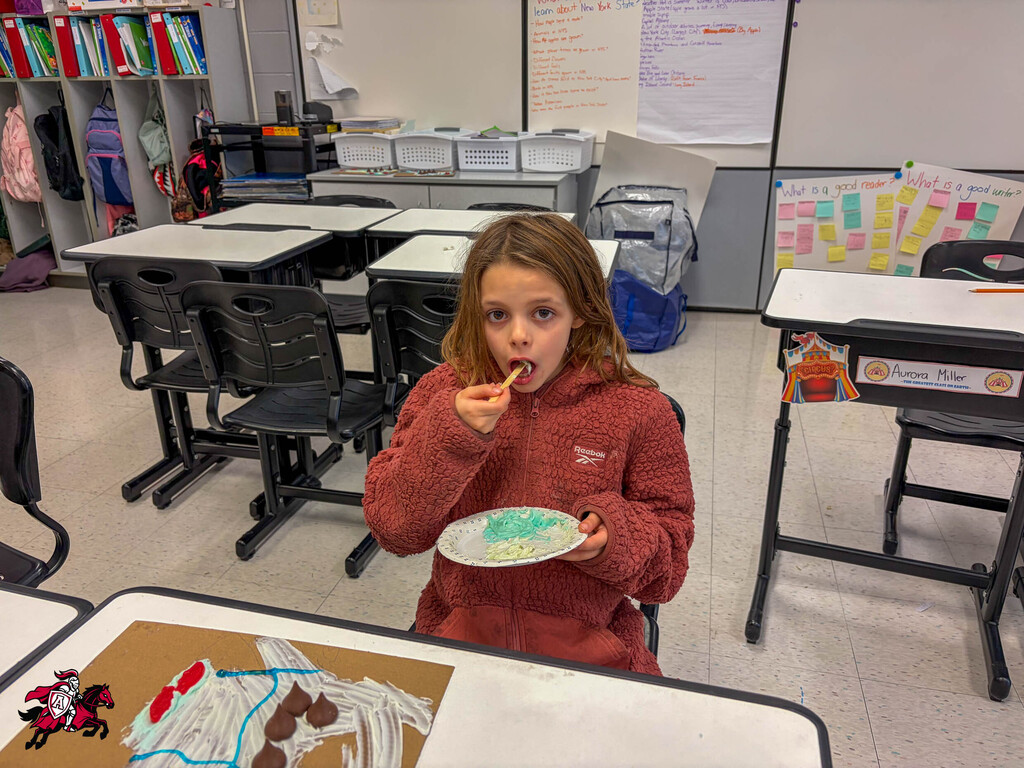A girl in a red sweater eats from a paper plate in a classroom with desks and chairs.