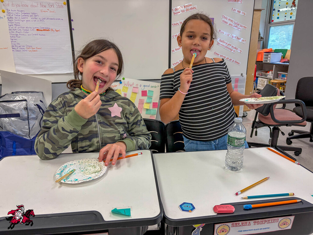 Two children in a classroom, one with a plate of food, the other holding a plate and a water bottle.