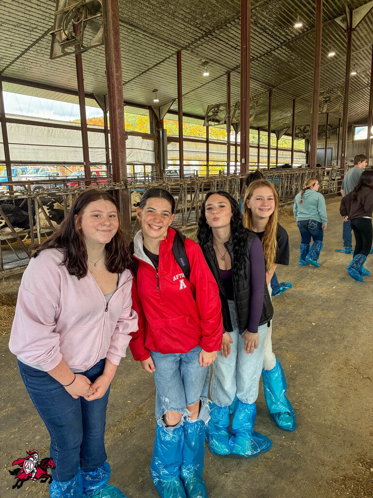 Five girls in boots stand together in a barn, one wearing a red jacket, another in pink. Behind them, metal beams and windows.