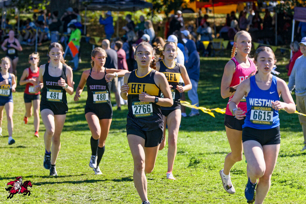 Runners compete in a cross-country race on a grassy field, with spectators watching from the sidelines.