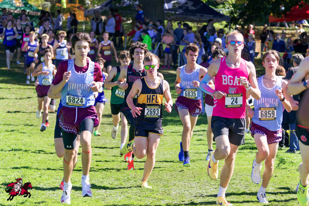 A group of runners compete in a cross-country race on a grassy field with a crowd in the background.