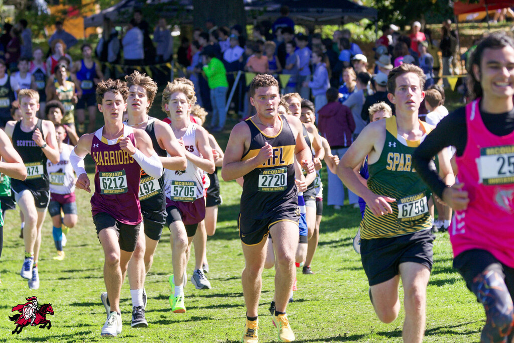 Runners compete in a cross-country race on a grassy field, with spectators in the background.