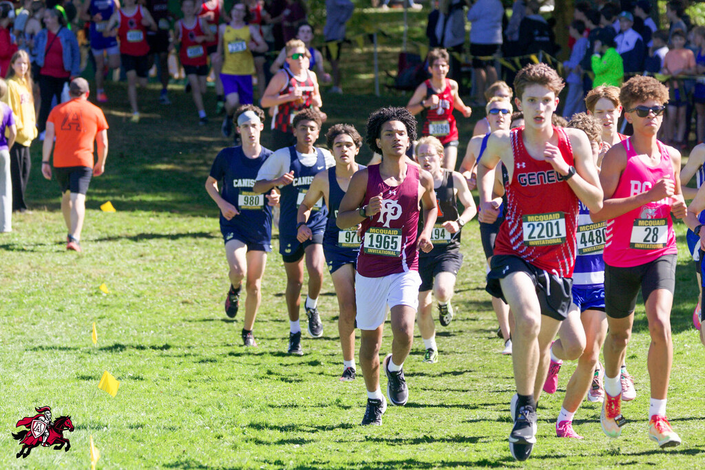 Runners compete in a cross-country race on a grassy field with spectators. Numbered bibs are visible.