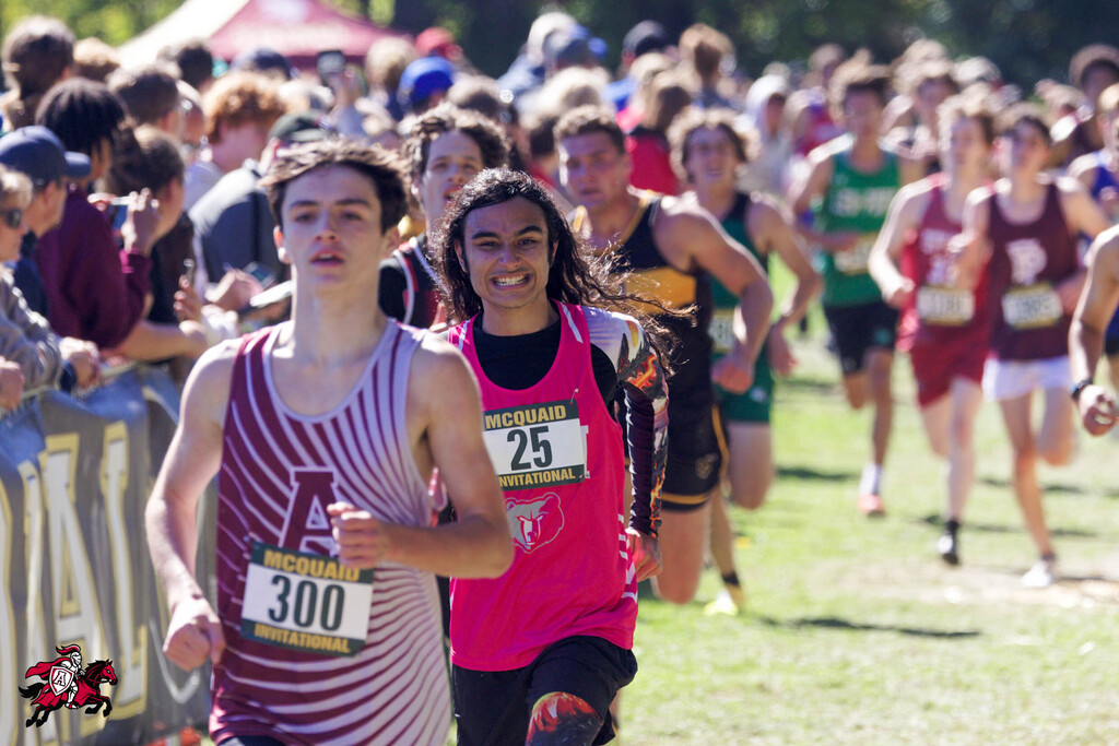 People running in a race on a grassy field. Participants wear numbered shirts, with one man in white and red and another in pink.