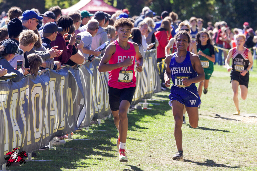 Several runners compete on a grassy field, with spectators behind a fence, some holding phones.