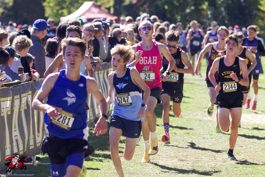 Runners compete in a cross-country race on a grassy field with spectators behind them.