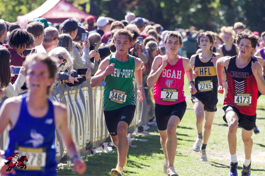 Runners in a race, wearing shirts with numbers, compete on grass. Spectators watch from behind a fence.