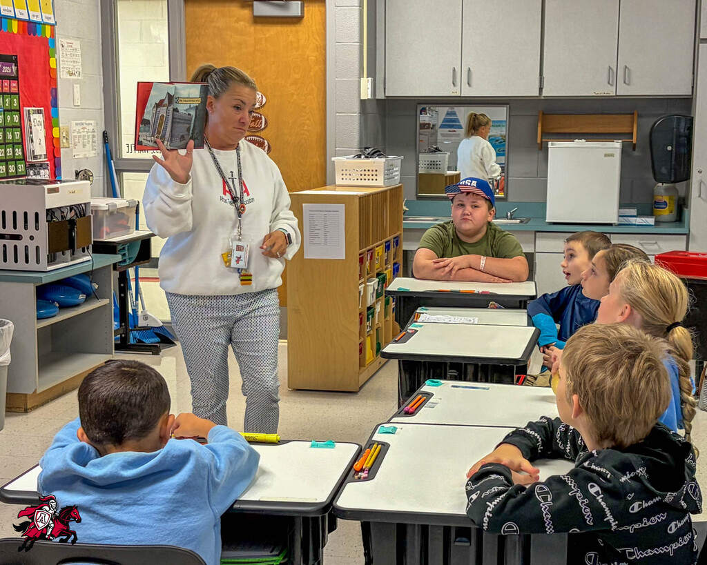 A woman stands in a classroom, holding a book, while children sit at desks, paying attention.