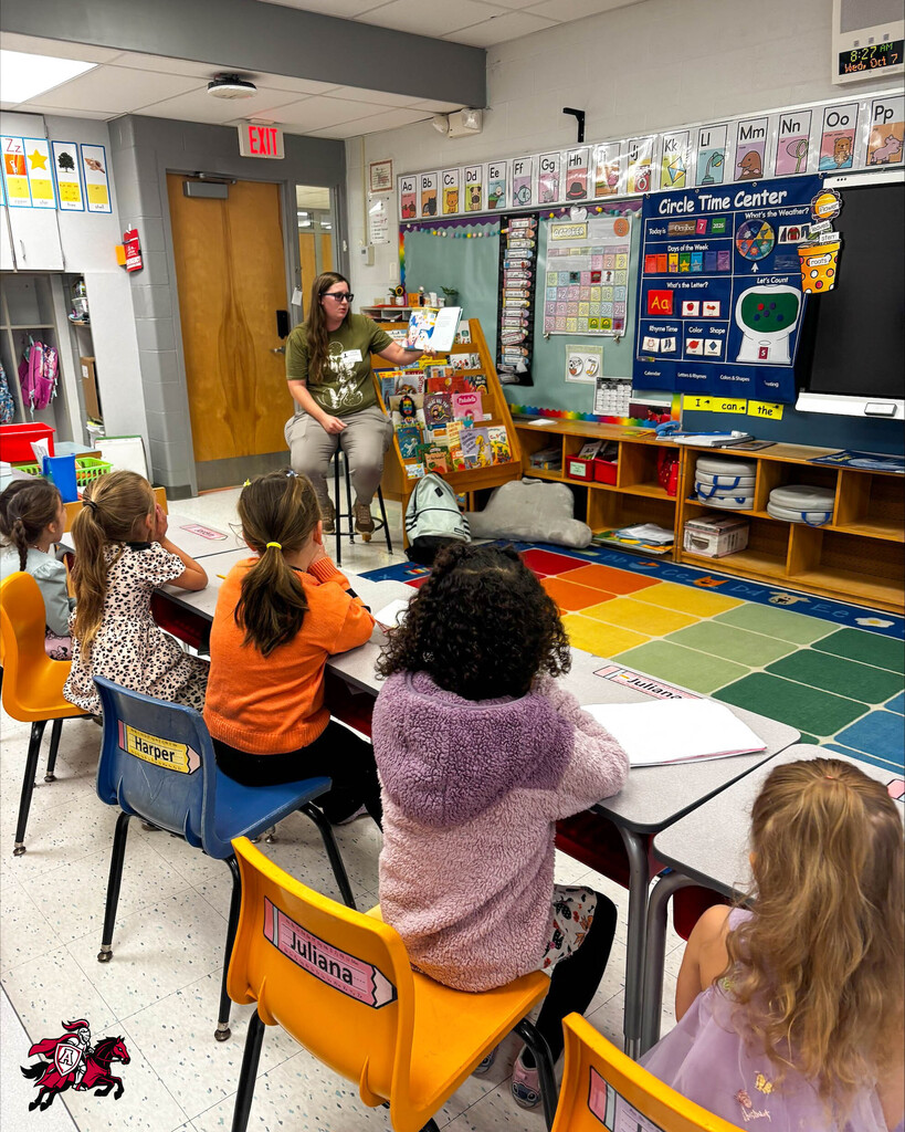 A teacher sits at a stool in front of children at desks, reading a book. Colorful rug on floor.