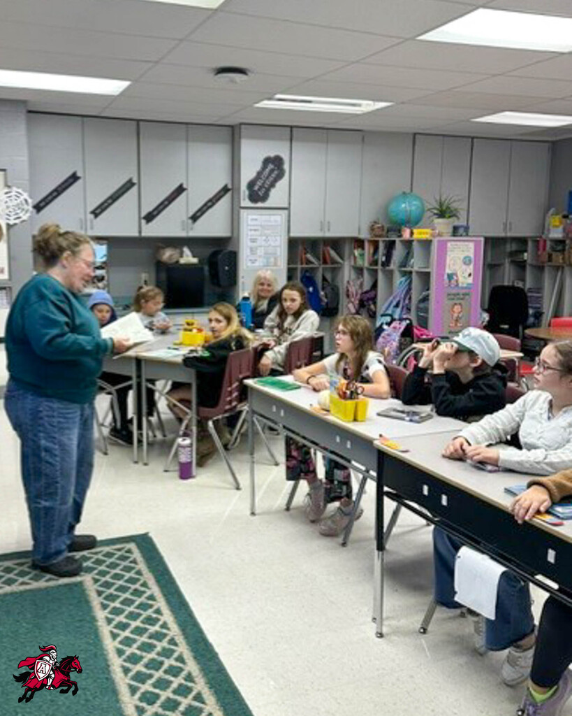 A teacher in a green sweater stands before a classroom with students seated at desks. Some students wear glasses.