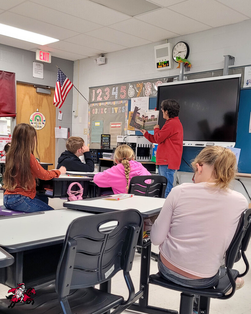 In a classroom, a person in a red shirt reads from a book while students in various shirts look on.