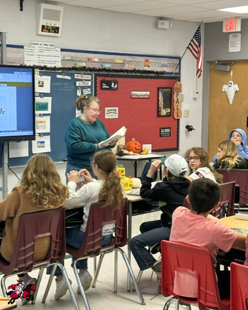 A classroom with students seated at desks, a teacher stands reading a book, and a monitor is on a wall.