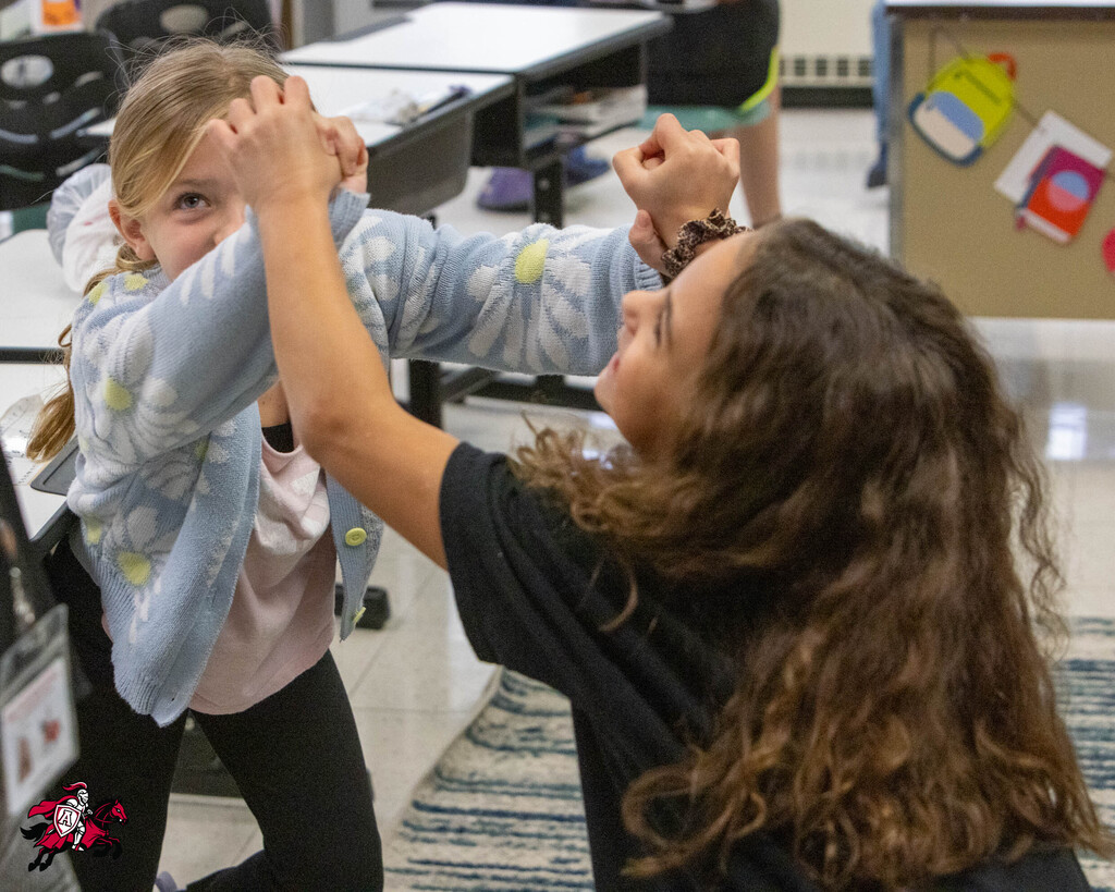 Two young girls playing in a classroom, one with curly hair and the other in a pink shirt.