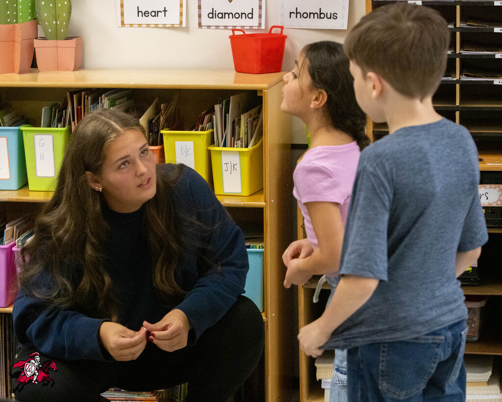 Woman crouches beside a boy and girl with books in a classroom. Behind them, shelves with labeled bins and potted plants.