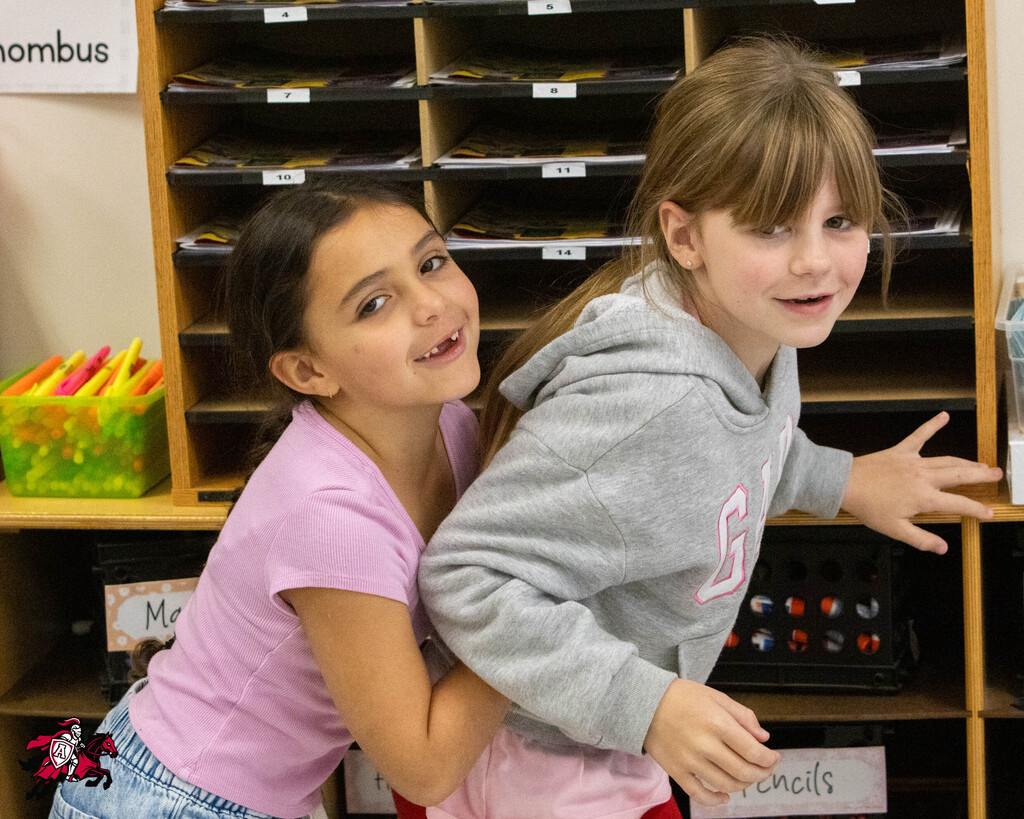 Two young girls in a classroom. One in pink, the other in gray. They are smiling and hugging.