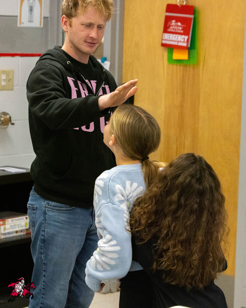 Man in black hoodie gestures while two girls, one with a ponytail, look at him in a room.