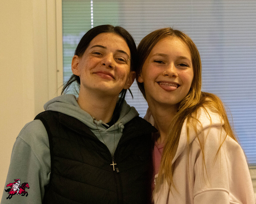 Two girls smile and stand together. One wears a gray hoodie with a black vest, the other a pink jacket. A white wall and window are behind them.