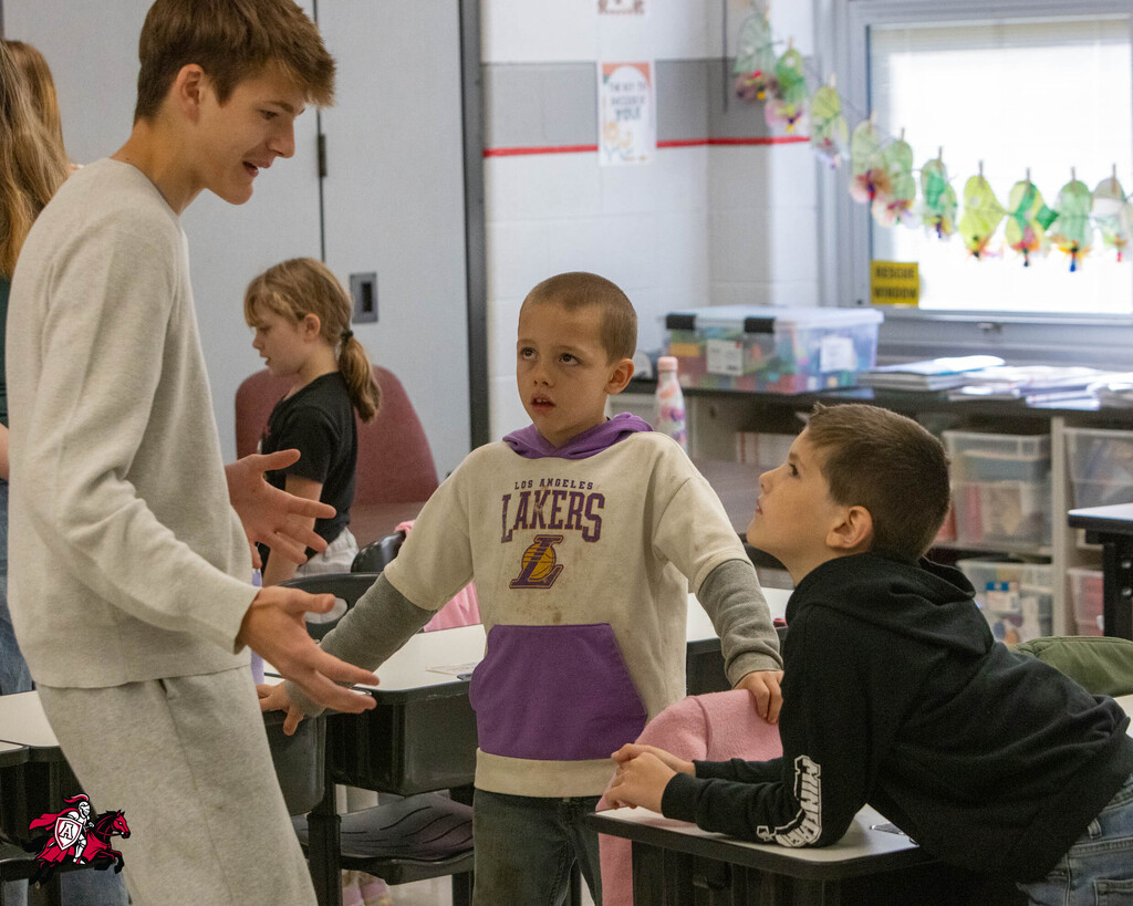 Three children in a classroom. One boy looks up at another boy in a Lakers sweatshirt while a third boy smiles at them.