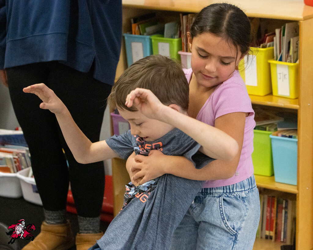 A girl holds a boy close to her chest in a classroom. Behind them, a wooden bookshelf filled with colorful bins and books.