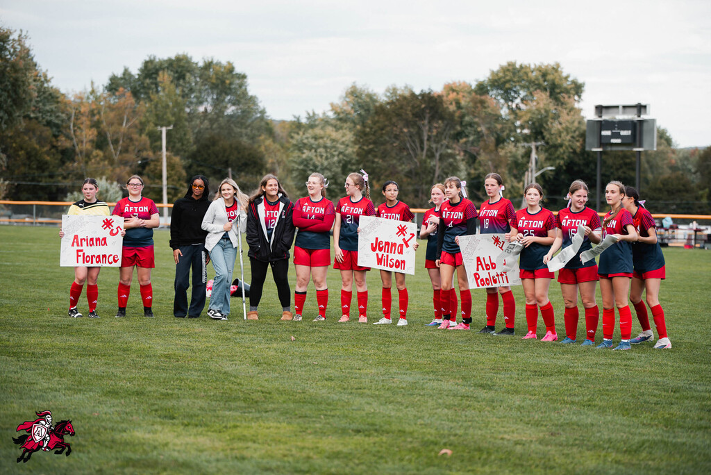 A team of female soccer players wearing red and black uniforms pose on a field with signs.