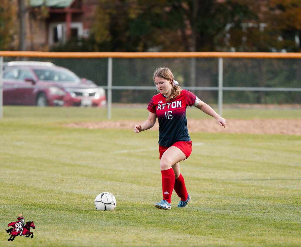 A woman in a red and black uniform kicks a soccer ball on a field with a fence.