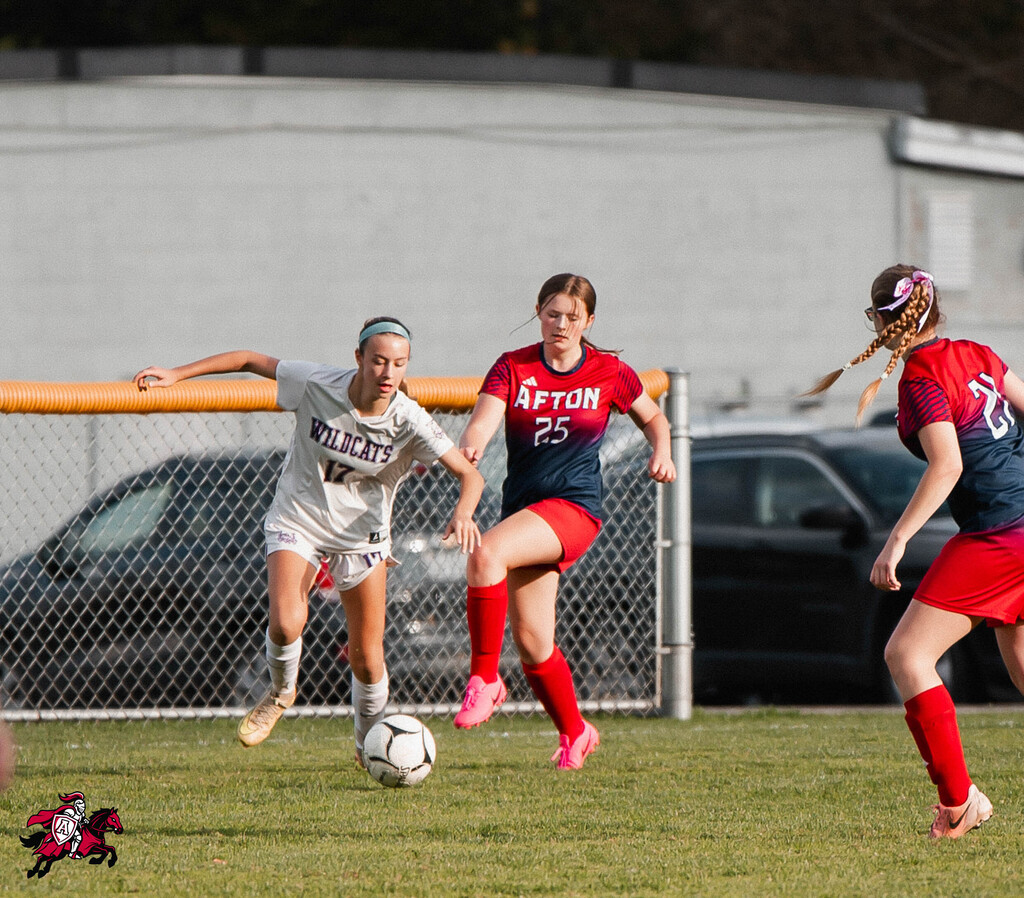 Three female soccer players on a field, two in red and one in white, with a soccer ball in the middle.