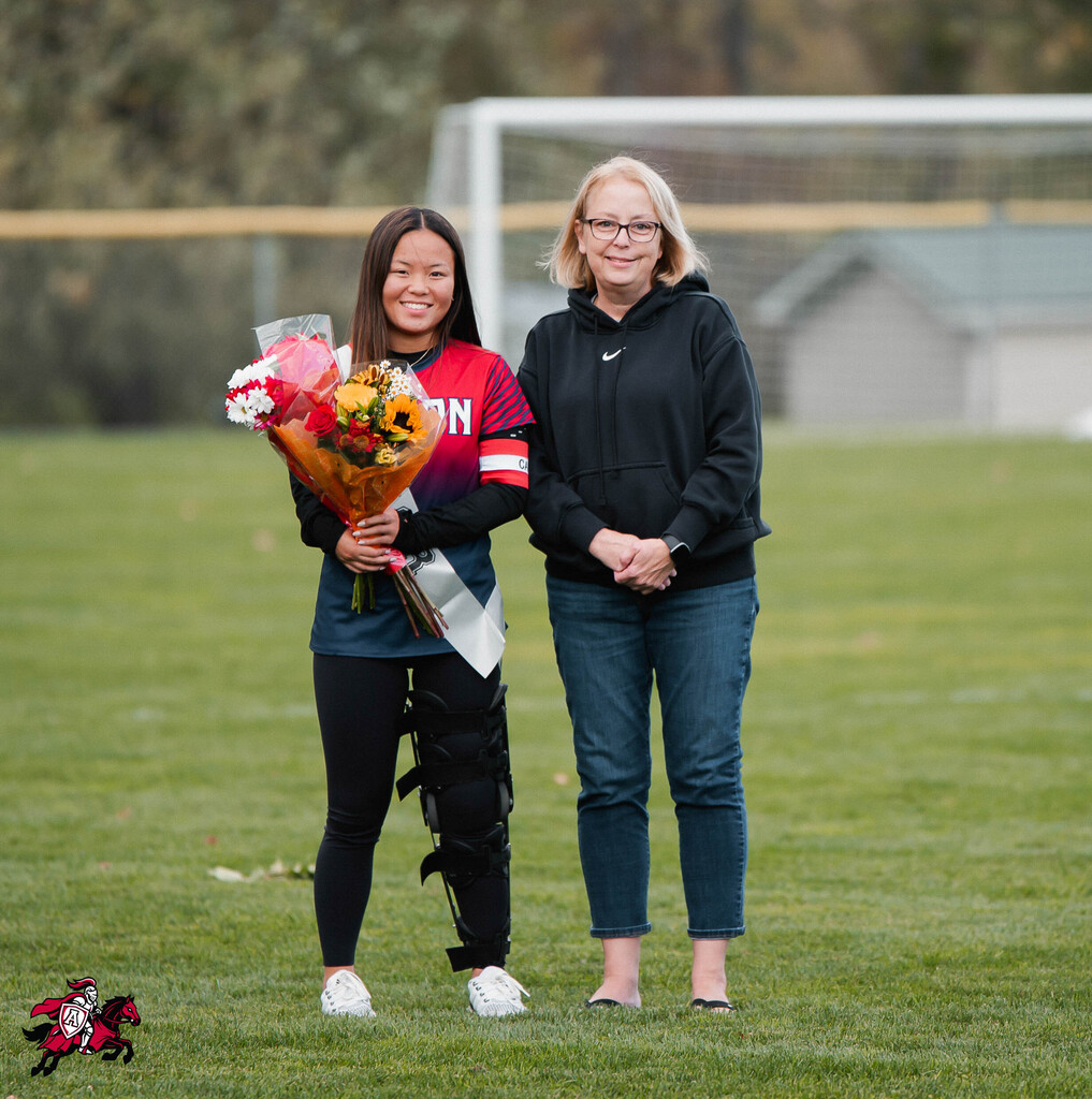 A young woman in a team jersey holds flowers. A woman stands beside her, both on a soccer field.