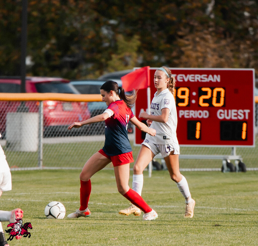 Two soccer players on a field with a red scoreboard. A red car is parked behind the fence.