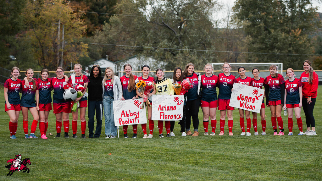 A team of female soccer players stands on a field with a coach holding signs, wearing red and blue uniforms.