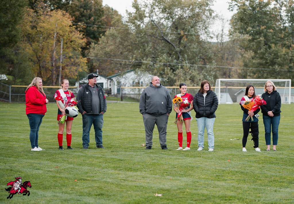A group of people stand on a grassy field, holding flowers. Some wear red shirts, others jackets. A soccer goal is behind them.