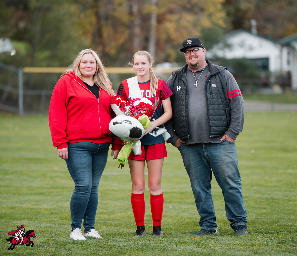 Three people on a soccer field: a woman in red, a girl in red shorts, and a man in gray.