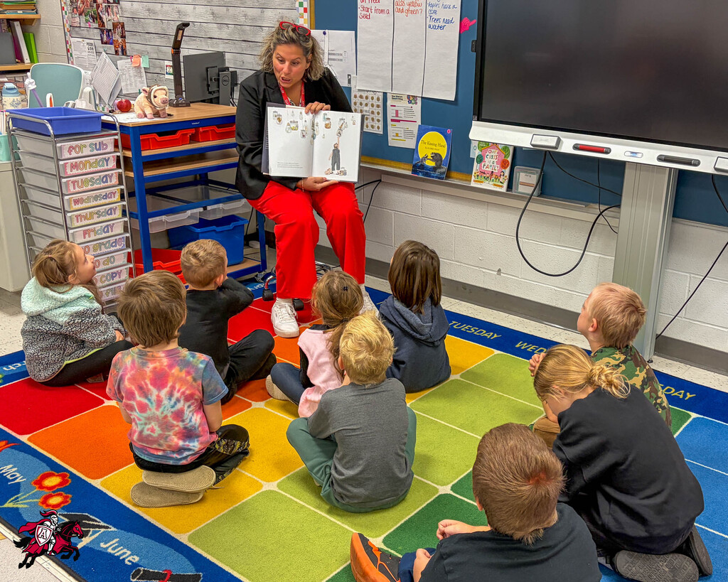 A woman in red pants reads a book to children seated in a circle on a colorful rug in a classroom.