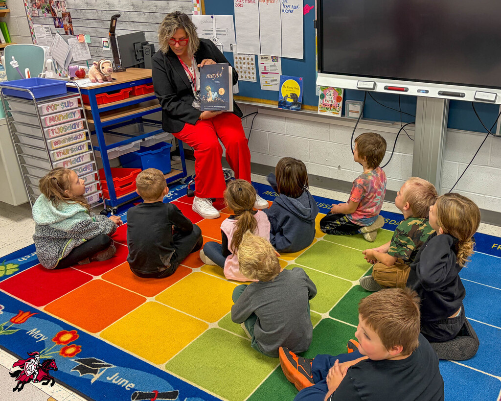 A teacher in red pants reads a book to a circle of young children sitting on a colorful rug in a classroom.