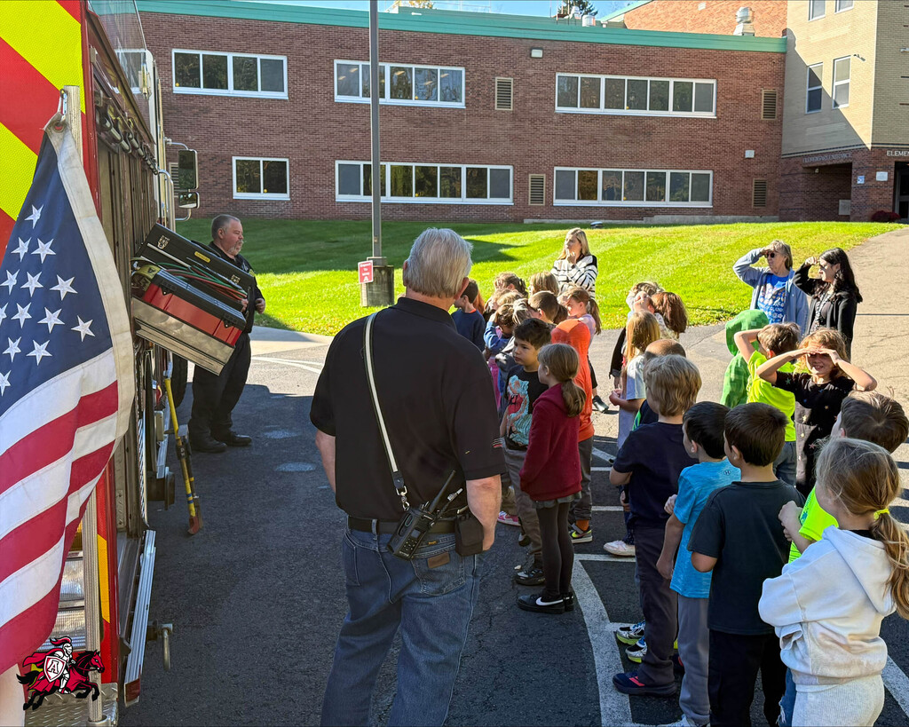 A line of children, some wearing vests, stand outdoors. A firefighter speaks to them while an American flag flies.