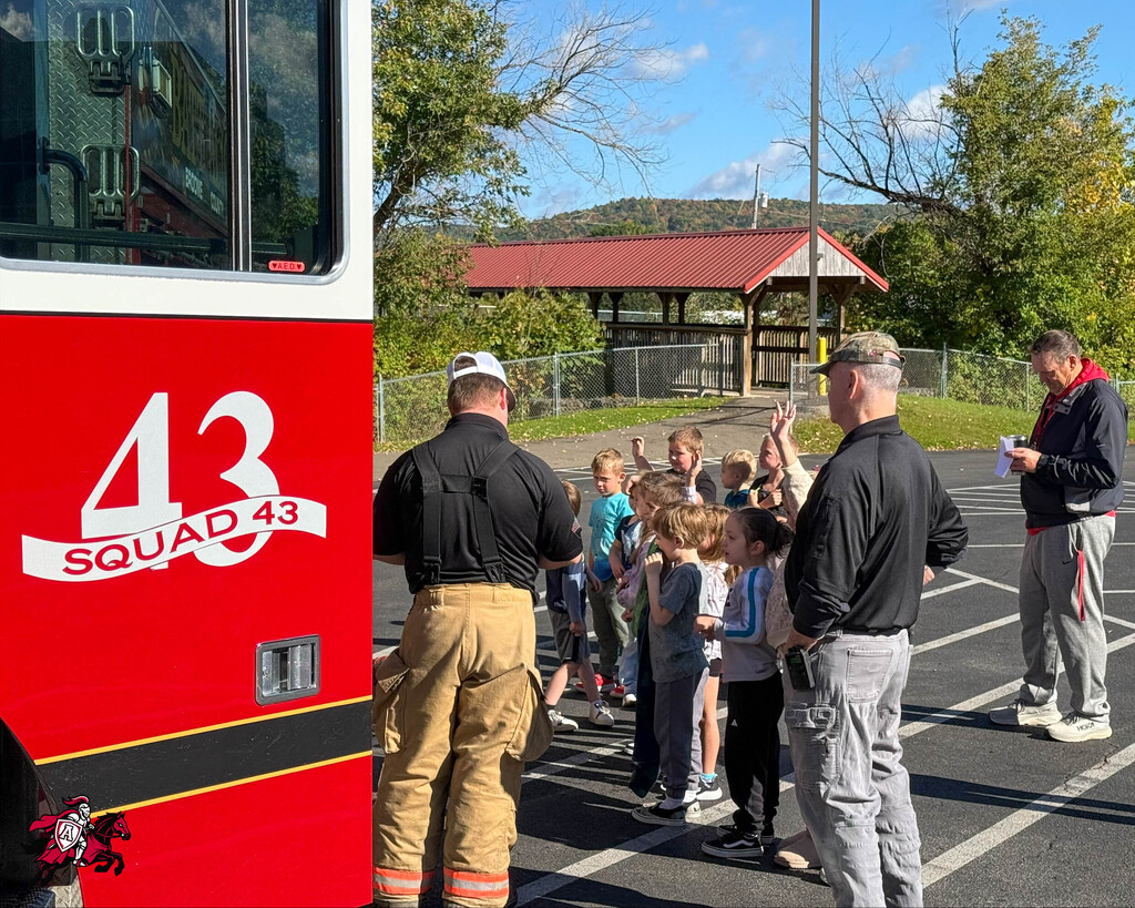 Firemen in uniform near a fire truck with a large crowd of children gathered in a parking lot.