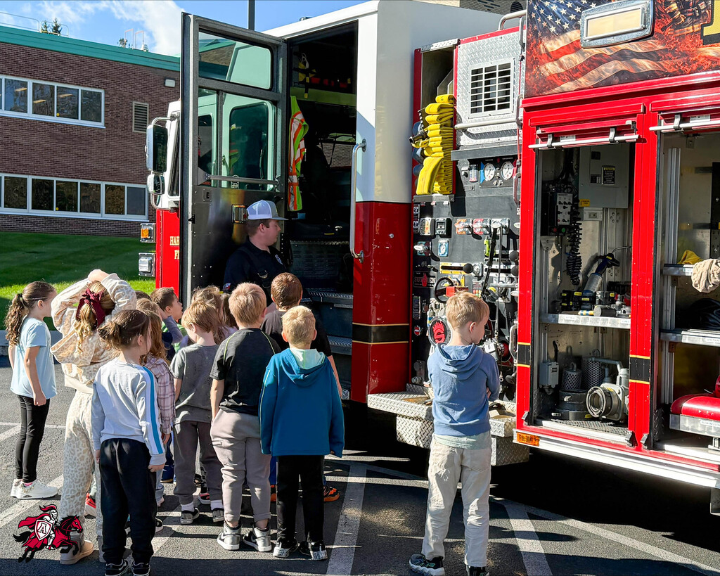 Children in front of a fire truck, with an adult standing inside, open doors, and fire equipment visible.