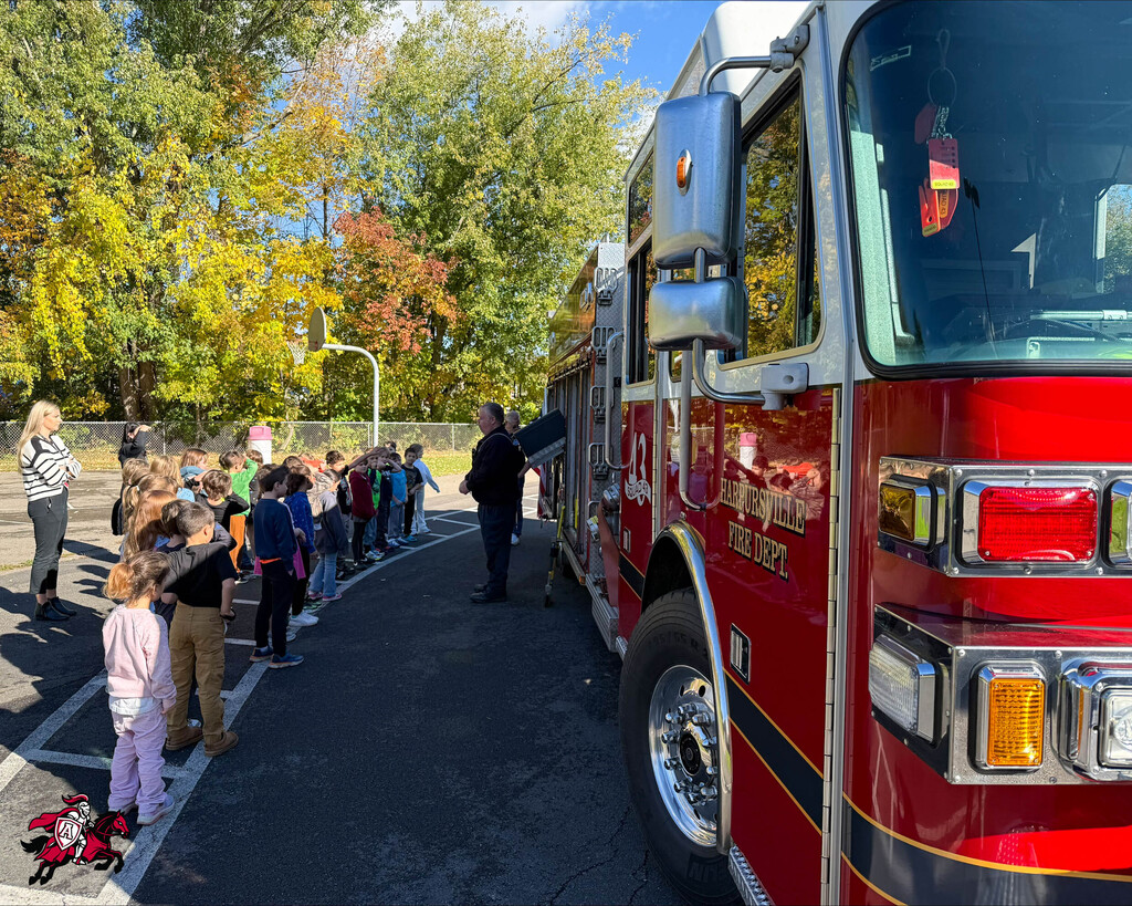 Line of children in front of a red fire truck. Trees and streetlight in the background.