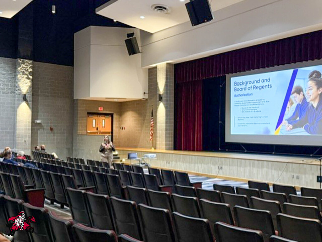 Empty auditorium with a stage. A person stands before a screen displaying a presentation. Red curtains hang in the background.