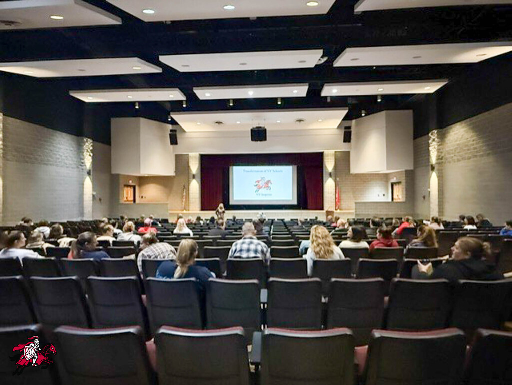 Auditorium with rows of seats, a large screen at the front, and several people seated in chairs.