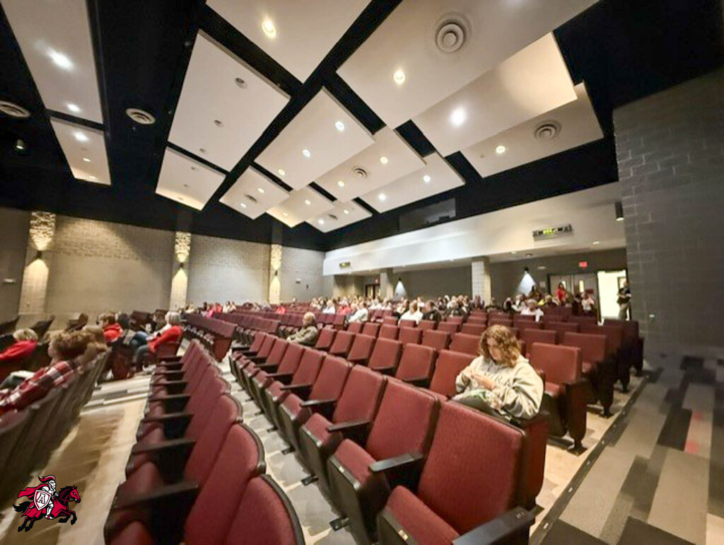 An empty auditorium with red chairs. A person sits in the center aisle. The ceiling has white panels and lights.