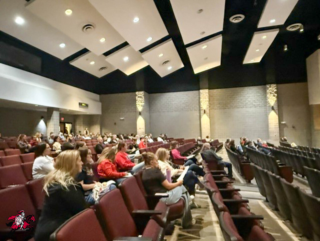An auditorium with rows of red chairs filled with people sitting, listening to a presentation.