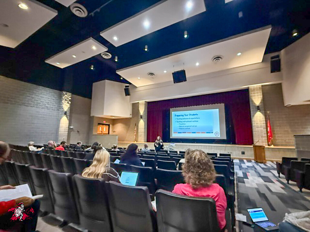 A room with rows of chairs, a person at the front with a projector, and people seated, looking at screens.