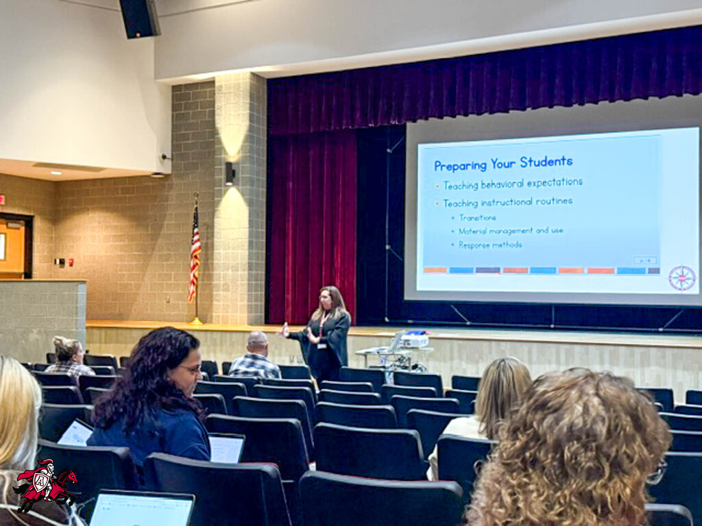 A woman presents to a seated audience in an auditorium with a projector screen displaying "Preparing Your Students" in the background.
