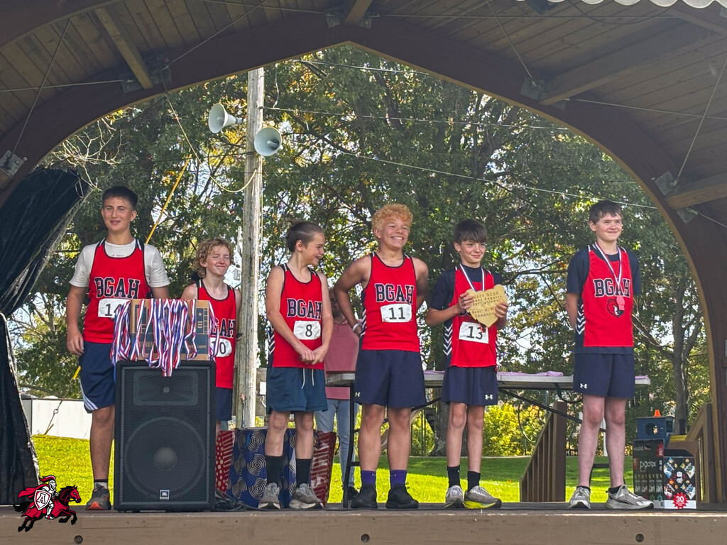 Six children stand on a stage, wearing matching red shirts, medals, and blue shorts. One holds a certificate.