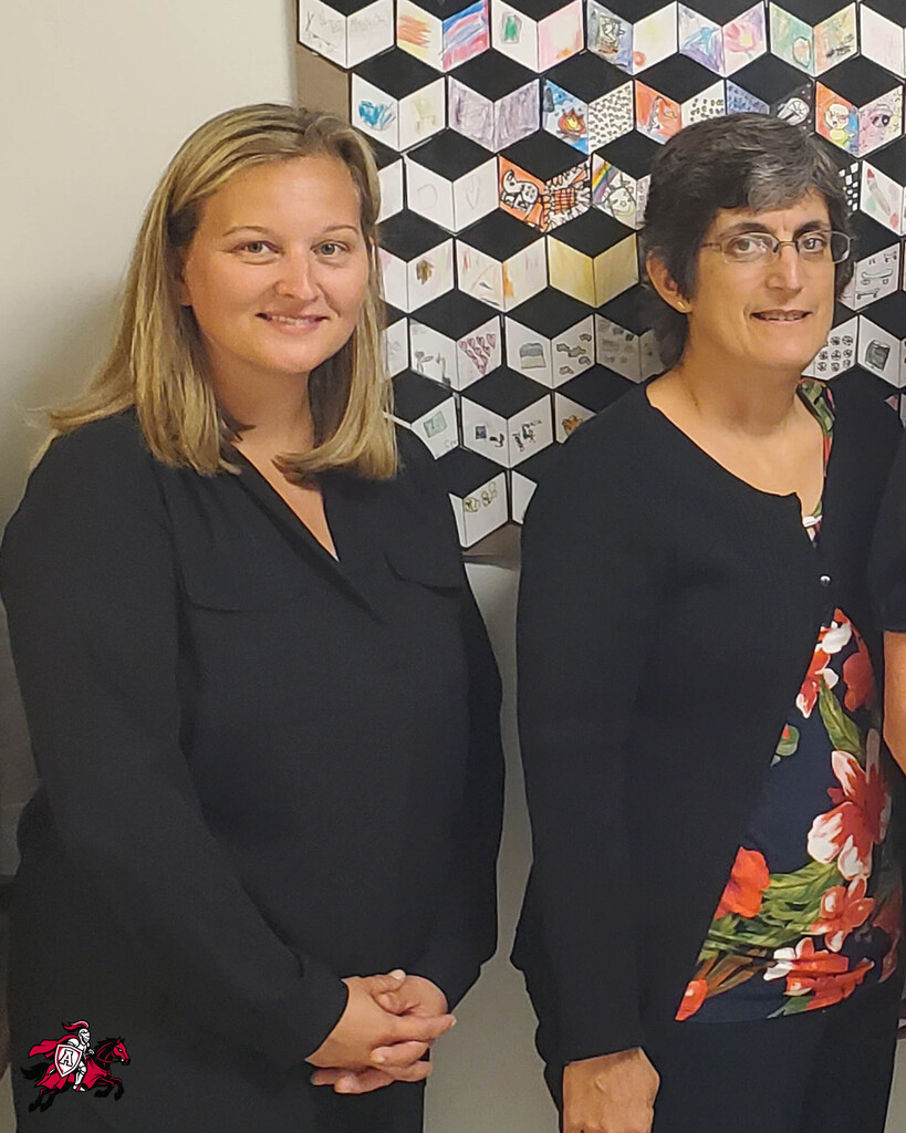 Two women stand beside each other; one with blonde hair, the other wearing glasses. They are in front of a colorful quilt.